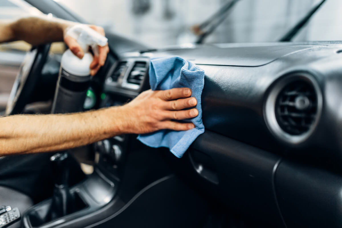 Hand reinigt das Armaturenbrett eines Autos mit Mikrofasertuch und Pflegespray